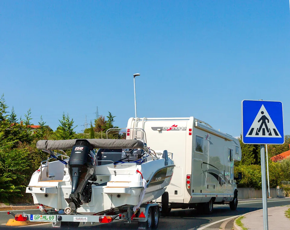 An RV pulls a boat along the road near a crosswalk, surrounded by trees and a clear sky.
