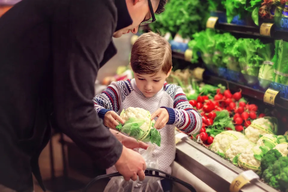 Adults and children pick out vegetables in the grocery store, focusing on a head of cauliflower