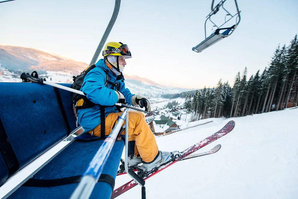 A skier on a ski lift, holding poles, looking out over a snowy mountain landscape, rocking outdoor recreation and the winter tourism economy.