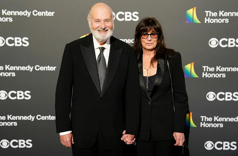 A couple poses on the red carpet at the Kennedy Center Honors event. The man is wearing a black suit and tie; the woman is wearing a black blazer and sunglasses