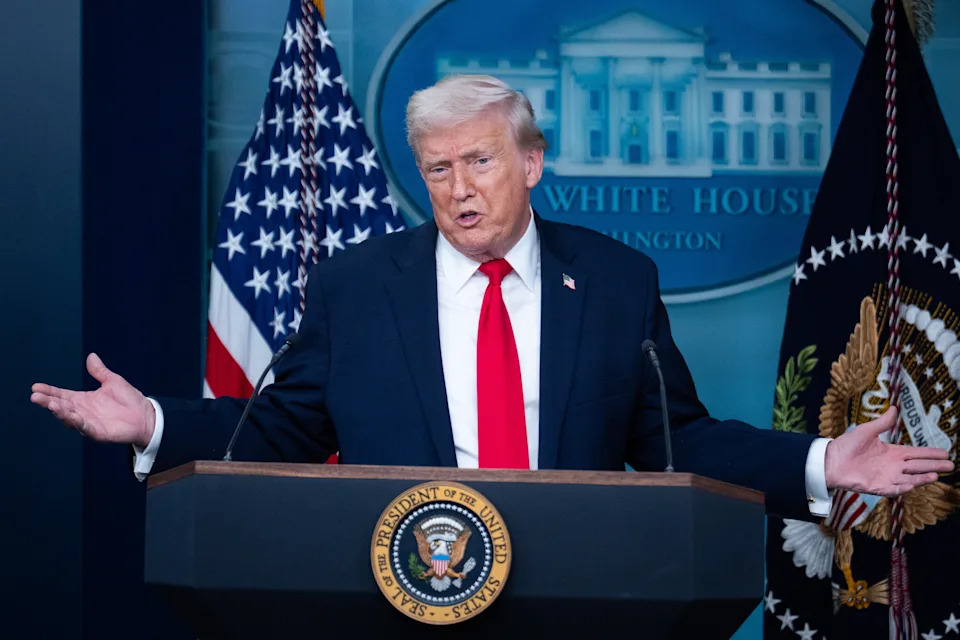 Person standing on stage with arms outstretched, sitting behind US flags and a blue sign.