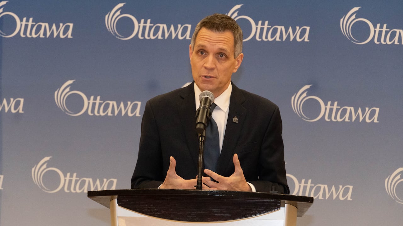 A man in a suit speaks on stage in front of a blue backdrop with the word Ottawa on it.