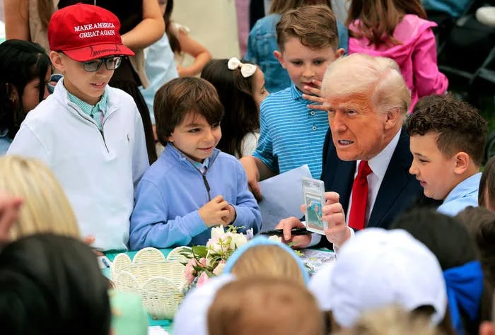A man in a suit is surrounded by children at a table with Easter items. One child is wearing red "Make America Great Again" hat
