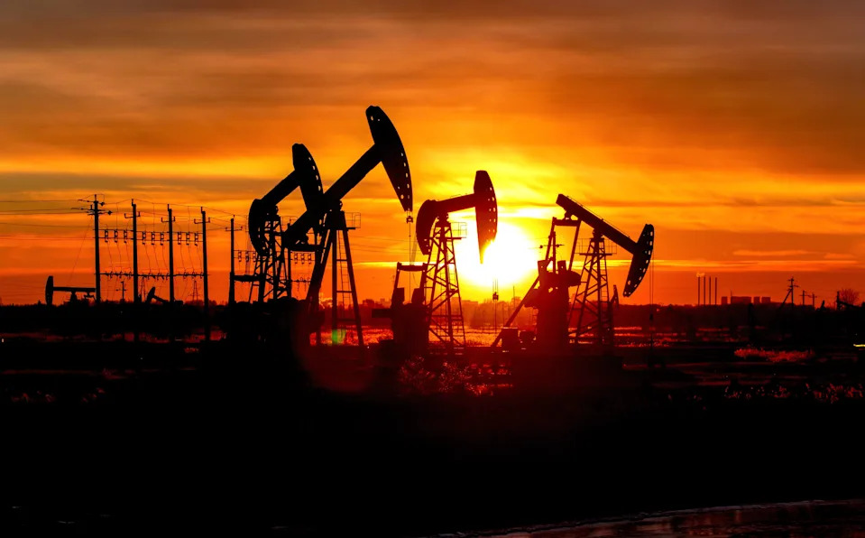 Oil pumps work against the setting sun, with towering figures and power lines in the background.
