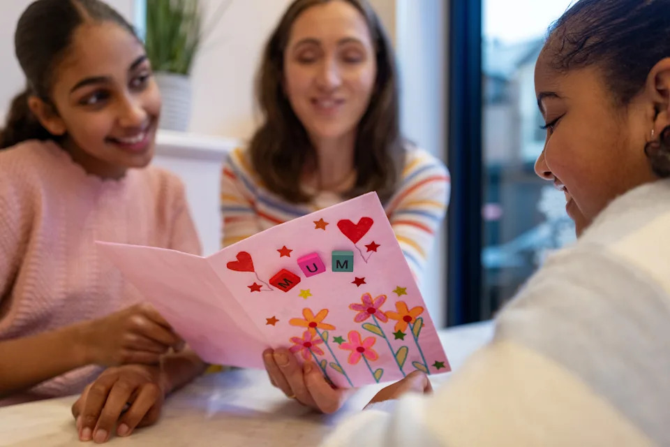 Three people smile as they look at a handmade, decorated card with a word on it "MOM" on it, with hearts and flowers