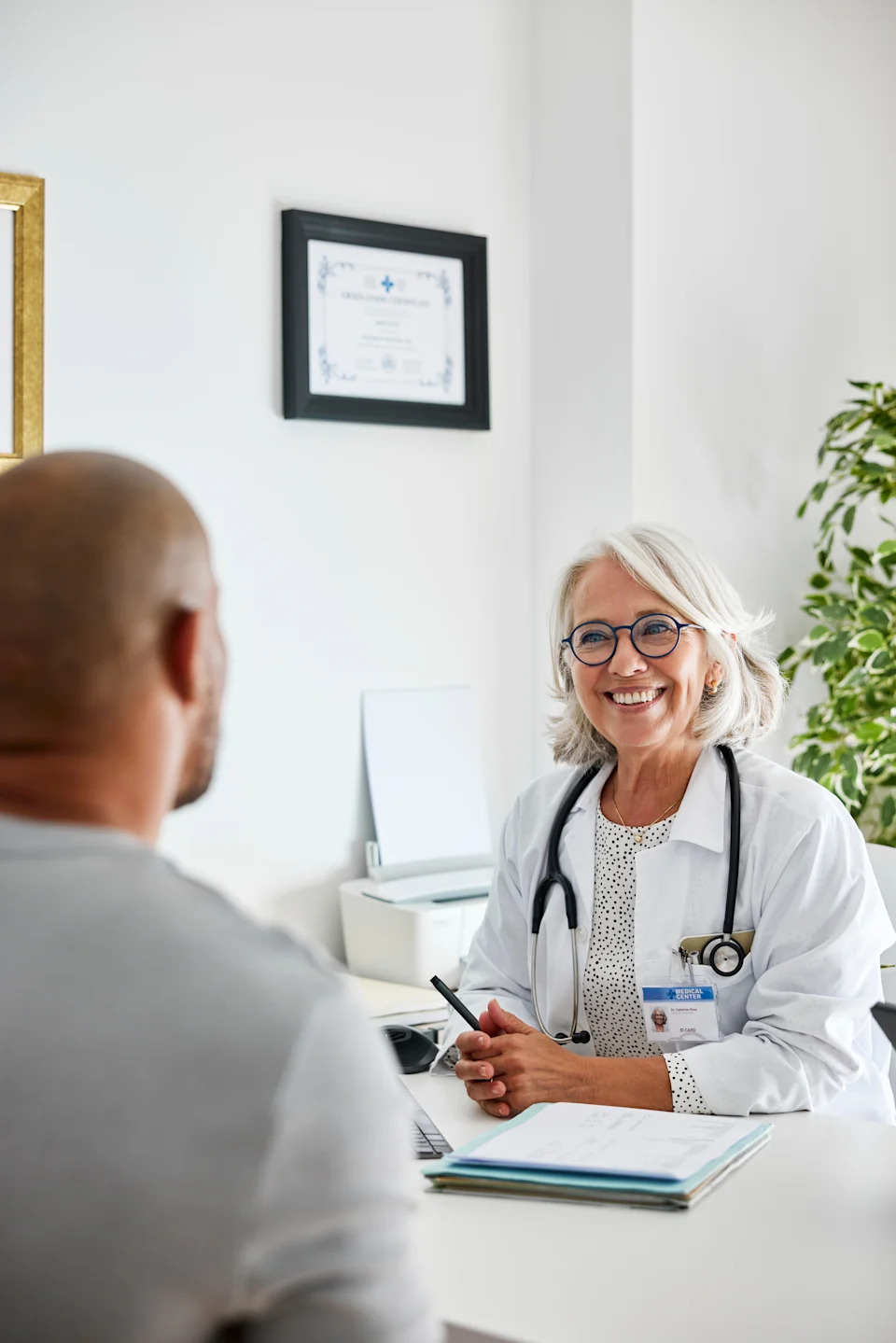 Smiling doctor with glasses, wearing lab coat and stethoscope, talking to patient in office space with certificate on wall