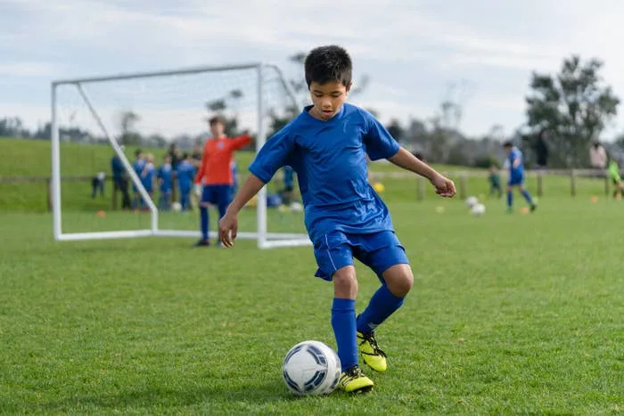 A boy in soccer jerseys knocks the ball down the field, with the goal and players behind